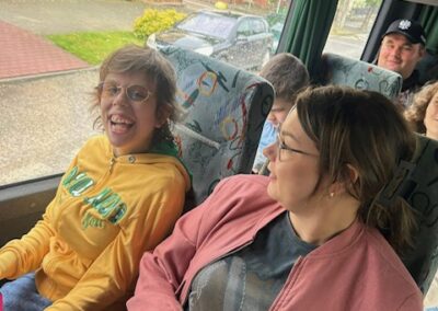 Dwóch pasażerów rozmawia w autobusie, na zewnątrz widać budynki i zaparkowane samochody.