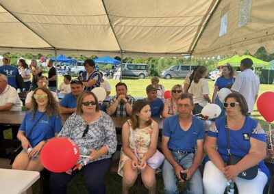 Ludzie siedzą pod namiotem na pikniku, obok stoi kilka samochodów i namiotów.