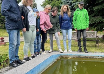 Siedem osób stoi nad oczkiem wodnym w parku, w tle drzewa i ławka, pochmurna pogoda.