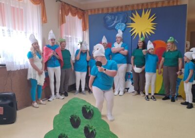 Grupa osób w kostiumach podobnych do Smerfów na tle kolorowej scenografii z dużym słoneczkiem i zielonym krzewem.