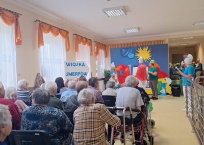 Starsze osoby oglądają przedstawienie teatralne o tematyce Smerfów w świetlicy.