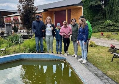 Sześć osób stoi przy oczku wodnym z liliami wodnymi w ogrodzie z drewnianym budynkiem w tle
