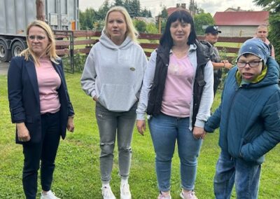 Grupa trzech kobiet i chłopiec stojący na trawniku na zewnątrz z ogrodzeniem i ciężarówką w tle.