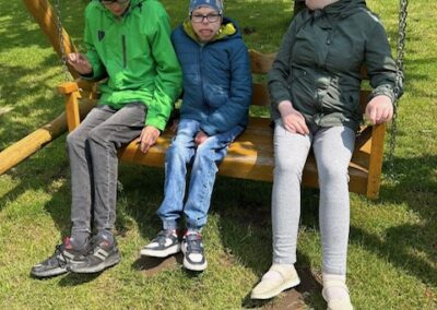 Trzy osoby siedzące na huśtawce drewnianej w parku na tle placu zabaw i trawnika.