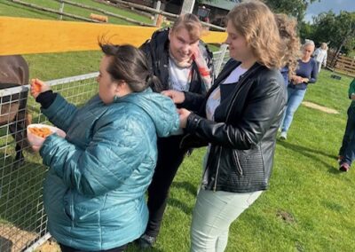 Trzy osoby karmią zwierzęta przez ogrodzenie na zielonym terenie z drzewami w tle.