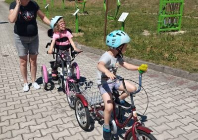 Chłopiec skręca rowerkiem na alejce, obok jedzie dziewczynka na trójkołowym rowerze, a opiekunka towarzyszy im podczas przejażdżki.