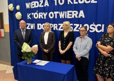 Osoby prowadzące stoją obok stołu z dokumentami i kwiatami przed kurtyną z hasłem Wiedza to klucz, który otwiera drzwi do przyszłości.