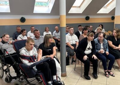 Uczestnicy siedzą na krzesłach w sali ze świetlikami w dachu, z przodu widoczna osoba na wózku oraz opiekunowie.