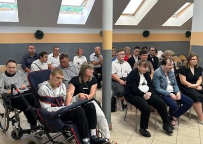 Widok na zebranych w sali z centralnym słupem i oknami dachowymi, w pierwszym rzędzie osoba na wózku i grupa siedzących osób.