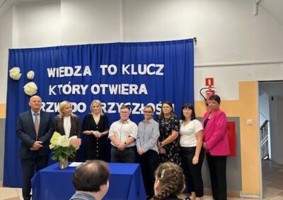 Grupa uczestników stoi przy stole z dokumentami i bukietem kwiatów na tle niebieskiej kurtyny z napisem Wiedza to klucz, który otwiera drzwi do przyszłości.