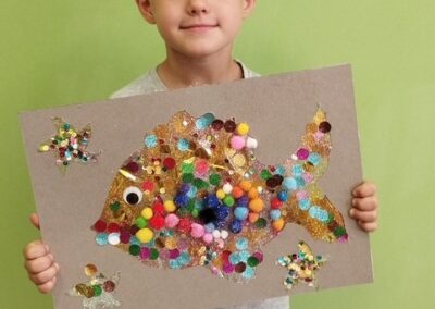Dziecko trzyma kartkę z kolorową rybką wyklejoną pomponami i cekinami na tle zielonej ściany.