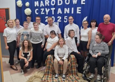 Grupa dzieci i dorosłych siedzi i stoi wspólnie przed granatową kurtyną z napisem Narodowe Czytanie 2025.