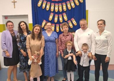 Grupa dzieci i nauczycieli pozująca do zdjęcia w udekorowanej sali podczas zakończenia roku przedszkolnego.