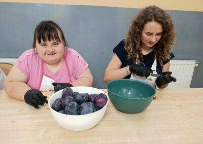 Dwie uczestniczki w fartuchach i rękawiczkach obierają śliwki przy stole, obok miska z owocami