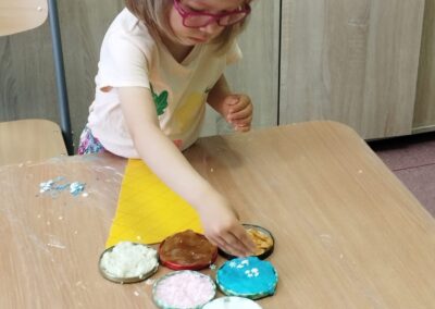 Dziewczynka w różowych okularach sięga po dekoracje z pojemników, przygotowując elementy do wyklejanki.