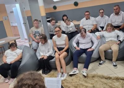 Uczestnicy siedzą na kanapie i krzesłach, słuchając czytanego tekstu w jasnej sali.