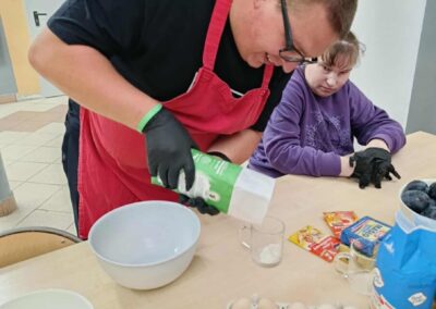 Uczestnik w czerwonym fartuchu wsypuje składnik do naczynia, na stole widoczne jajka, mąka i miski