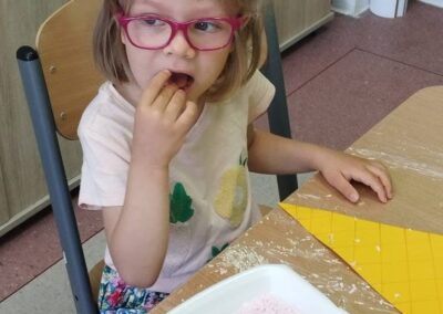 Dziewczynka w różowych okularach wkłada do ust przekąskę, obok leży taca z różowymi wiórkami i żółty papier do pracy.