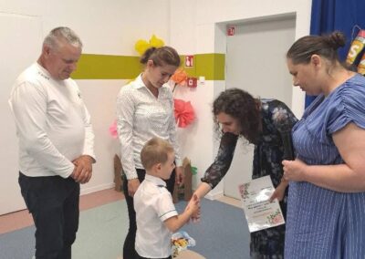 Dziecko odbiera dyplom od nauczyciela w obecności kadry podczas uroczystości zakończenia roku.