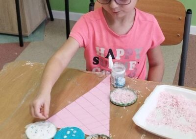 Dziewczynka układa ozdoby i kolorowe masy w zakrętkach, pracując nad rożkiem lodowym z papieru na stole.