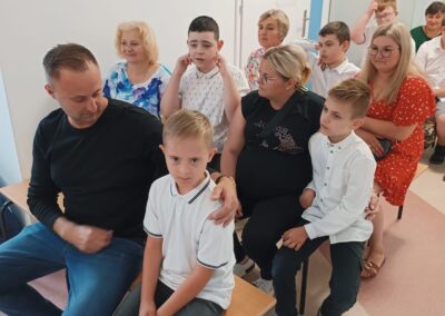 Publiczność złożona z dzieci i dorosłych siedzi na krzesłach podczas szkolnej uroczystości.