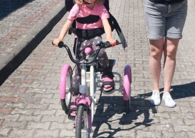 Dziewczynka w kasku jedzie na trójkołowym rowerze rehabilitacyjnym, a dorosła osoba idzie obok i pomaga.