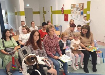Dzieci i dorośli siedzący na krzesłach w sali podczas uroczystości zakończenia roku przedszkolnego.