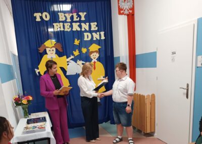 Nauczycielka wręcza uczniowi dyplom na tle dekoracji zakończenia roku.