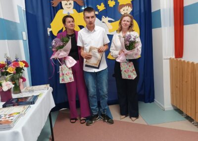 Uczeń z dyplomem stoi obok nauczycielek na tle szkolnej dekoracji.