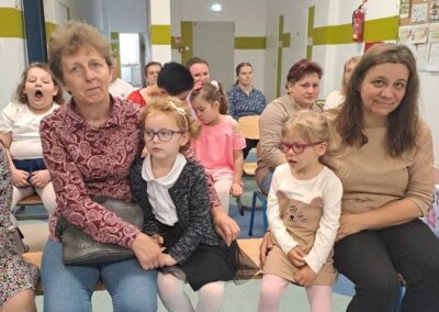 Zebrani dorośli i dzieci siedzący na widowni w sali przedszkolnej podczas uroczystości.