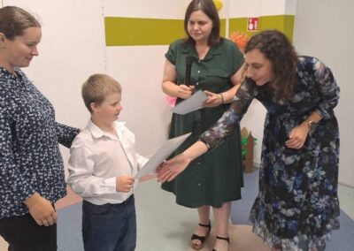 Nauczyciel wręcza dziecku dyplom w sali przedszkolnej; obok stoją inne osoby dorosłe.