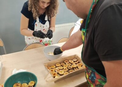 Uczestnik układa połówki śliwek na cieście w dużej blaszce, w tle druga osoba przygotowuje owoce