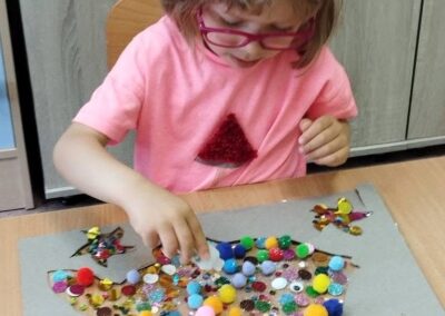 Dziewczynka w okularach przykleja pompony i błyszczące elementy do papierowej rybki ułożonej na szarym podkładzie.