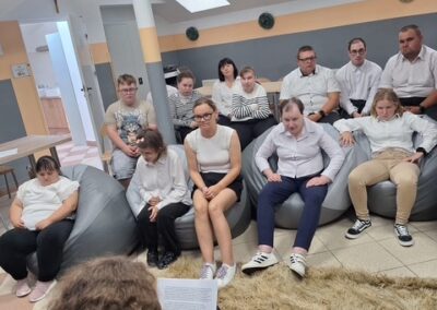 Uczestnicy siedzą w sali, w tle widać scenę i osobę czytającą.