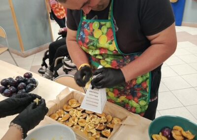 Uczestnik w fartuchu rozkłada połówki śliwek na cieście, obok miski z masą i owocami