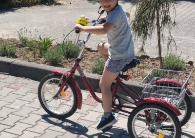 Chłopiec w jasnym kasku siedzi na czerwonym trójkołowym rowerku i spogląda w stronę aparatu na słonecznej alejce.