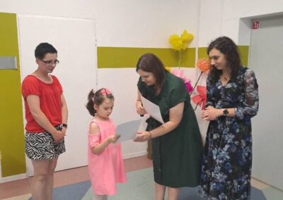 Dziecko odbiera dyplom od nauczyciela w sali przedszkolnej; obok stoją nauczyciele.