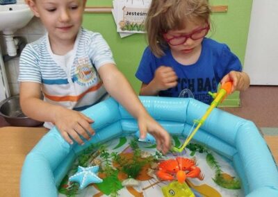 Dwoje dzieci bawi się przy dmuchanym baseniku z wodą, łowiąc zabawkowe rybki i kraba wędką magnetyczną.