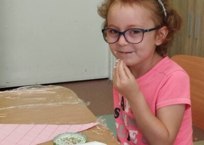 Dziewczynka w okularach trzyma w dłoni wiórki dekoracyjne i z uśmiechem próbuje ich przy stole z materiałami plastycznymi.