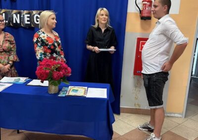 Nauczyciel wręcza uczniowi dyplom na tle szkolnej dekoracji zakończenia roku.