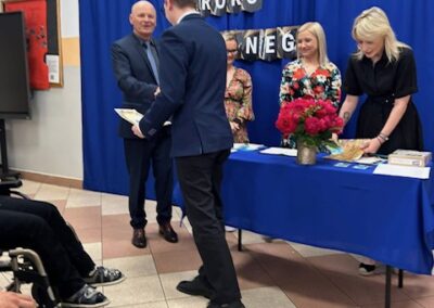 Uczeń stoi na scenie obok nauczycieli w trakcie wręczania nagrody podczas uroczystości szkolnej.