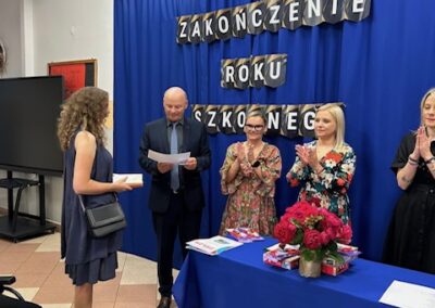 Uczniowie i nauczyciele na scenie w trakcie wręczania wyróżnień na zakończenie roku szkolnego.