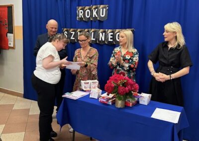 Uczeń z dyplomem stoi na scenie na tle dekoracji zakończenia roku szkolnego.